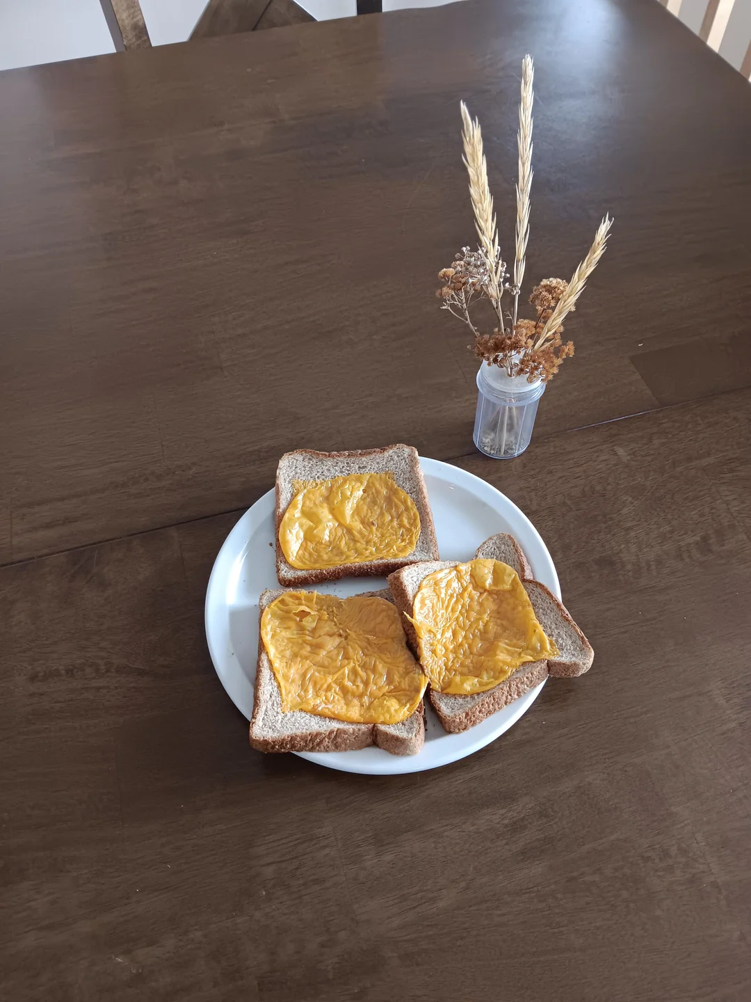 Three slices of wheat toast with melted yellow cheese on a white plate sit on a brown table next to a small vase holding dried grasses and flowers.