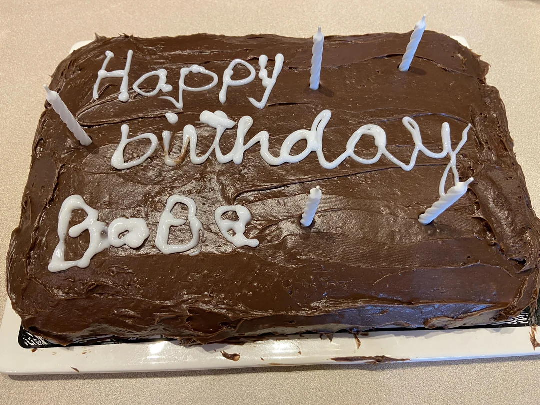 A rectangular chocolate cake with five unlit white candles and “Happy birthday Babe” written in white icing on top.