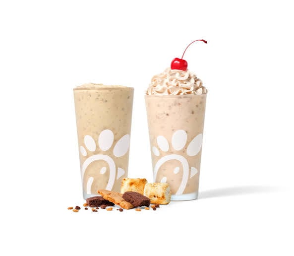 Two Chick-fil-A milkshakes, one plain and one topped with whipped cream and a cherry, are shown with crumbled chocolate cookies, graham crackers, and marshmallows in front.