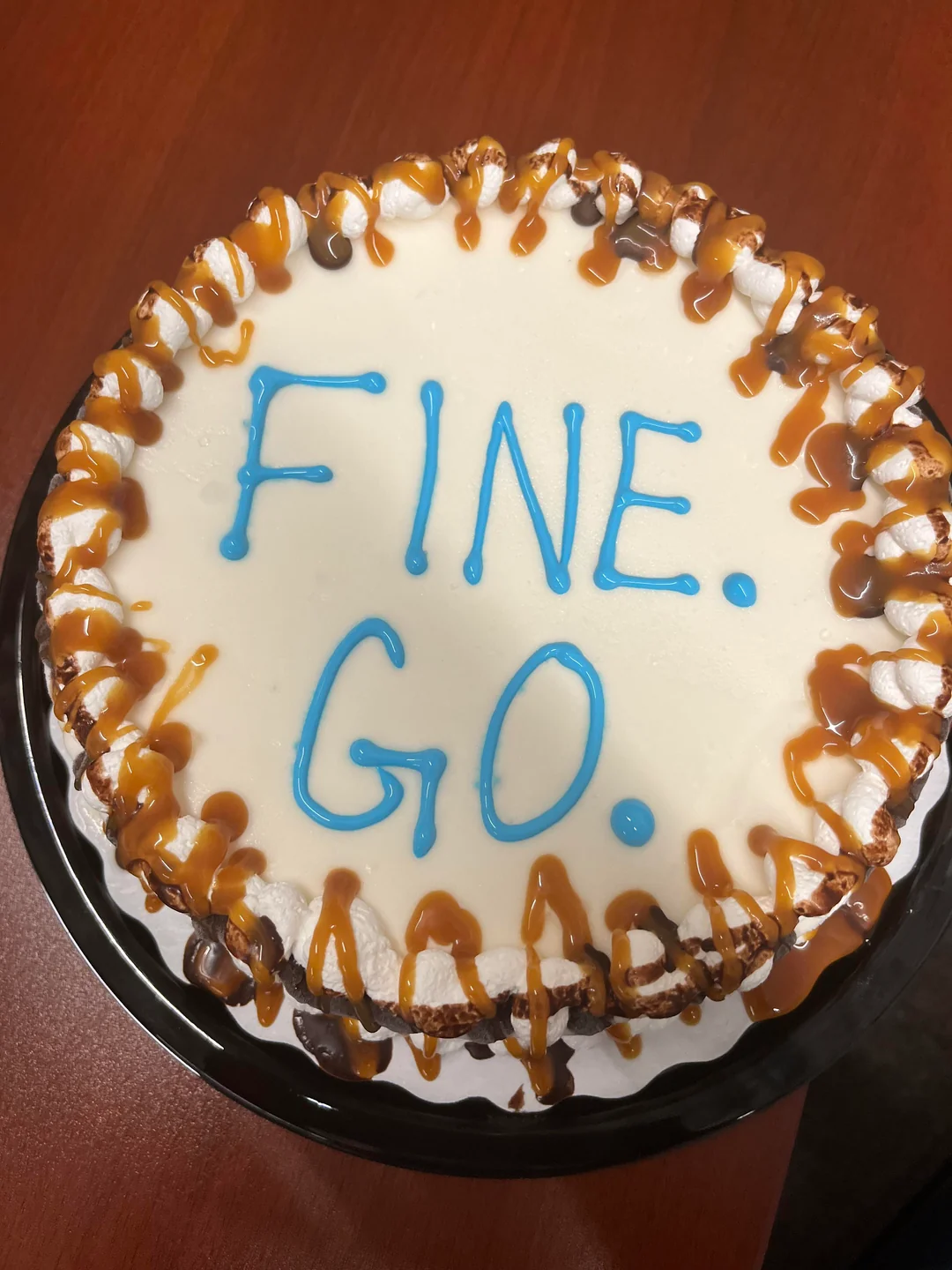 A round cake with white frosting, blue icing letters reading “FINE. GO.”, and caramel drizzle on the edge, sitting on a wooden table.