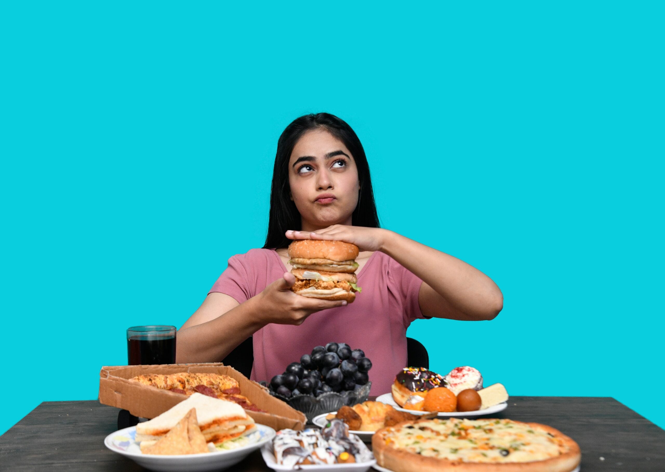 A woman sits at a table with various fast foods, holding a large burger with both hands and looking upward thoughtfully. The foods include pizza, donuts, grapes, a sandwich, and a drink, against a solid turquoise background.