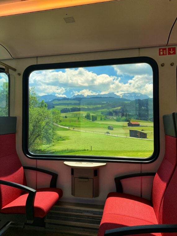 A train window frames a scenic view of green fields, hills, and distant mountains under a blue sky with clouds. Two red train seats and a small table are visible in the foreground.