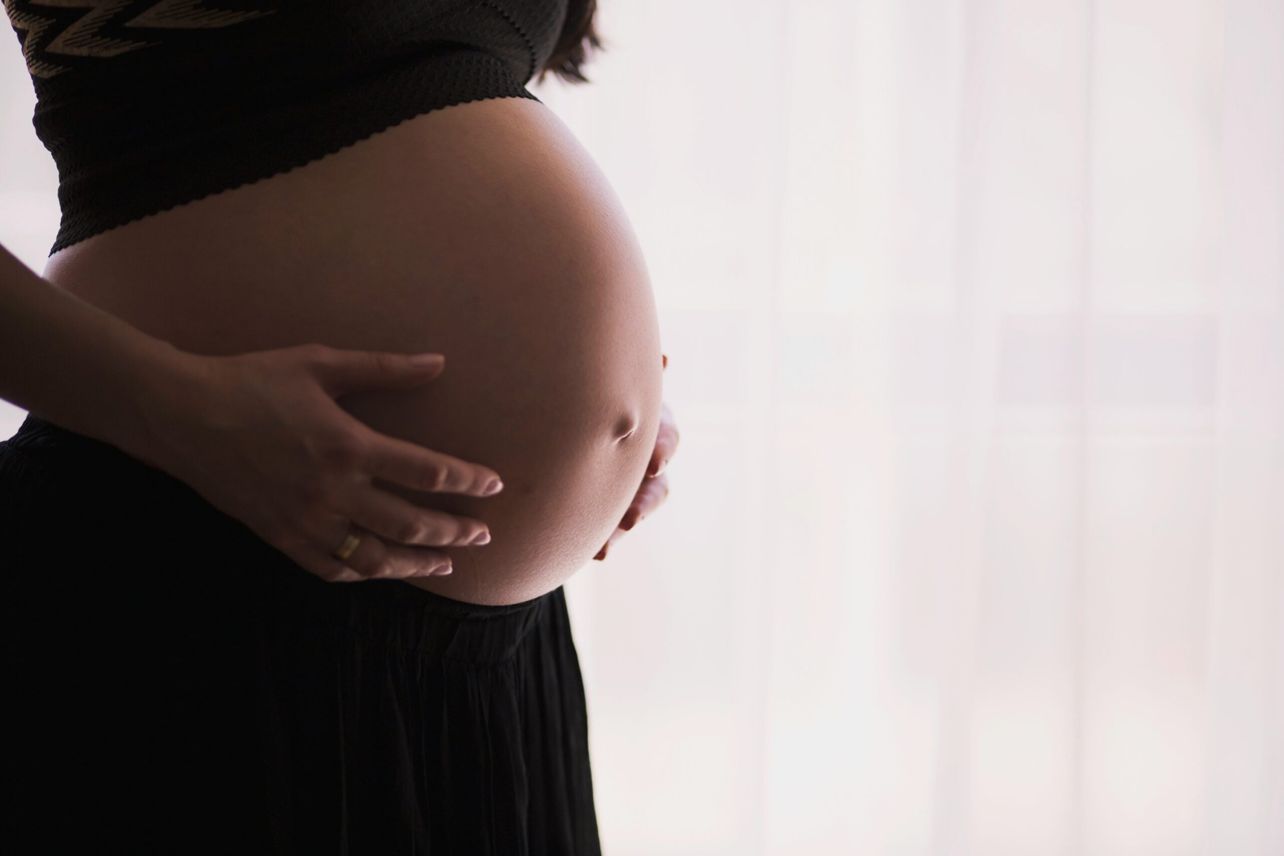 A person wearing a black skirt and top gently cradles their pregnant belly with both hands, standing in front of a softly lit window, quietly reflecting on the life in 2026 problems that await the next generation.