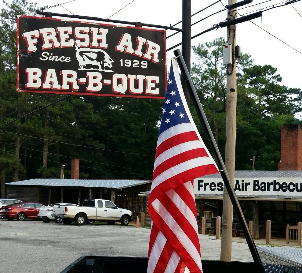 A U.S. flag is in front of a sign reading “Fresh Air Bar-B-Que Since 1929” with a pig logo. Behind are trees, parked cars, and a building labeled “Fresh Air Barbecue.”