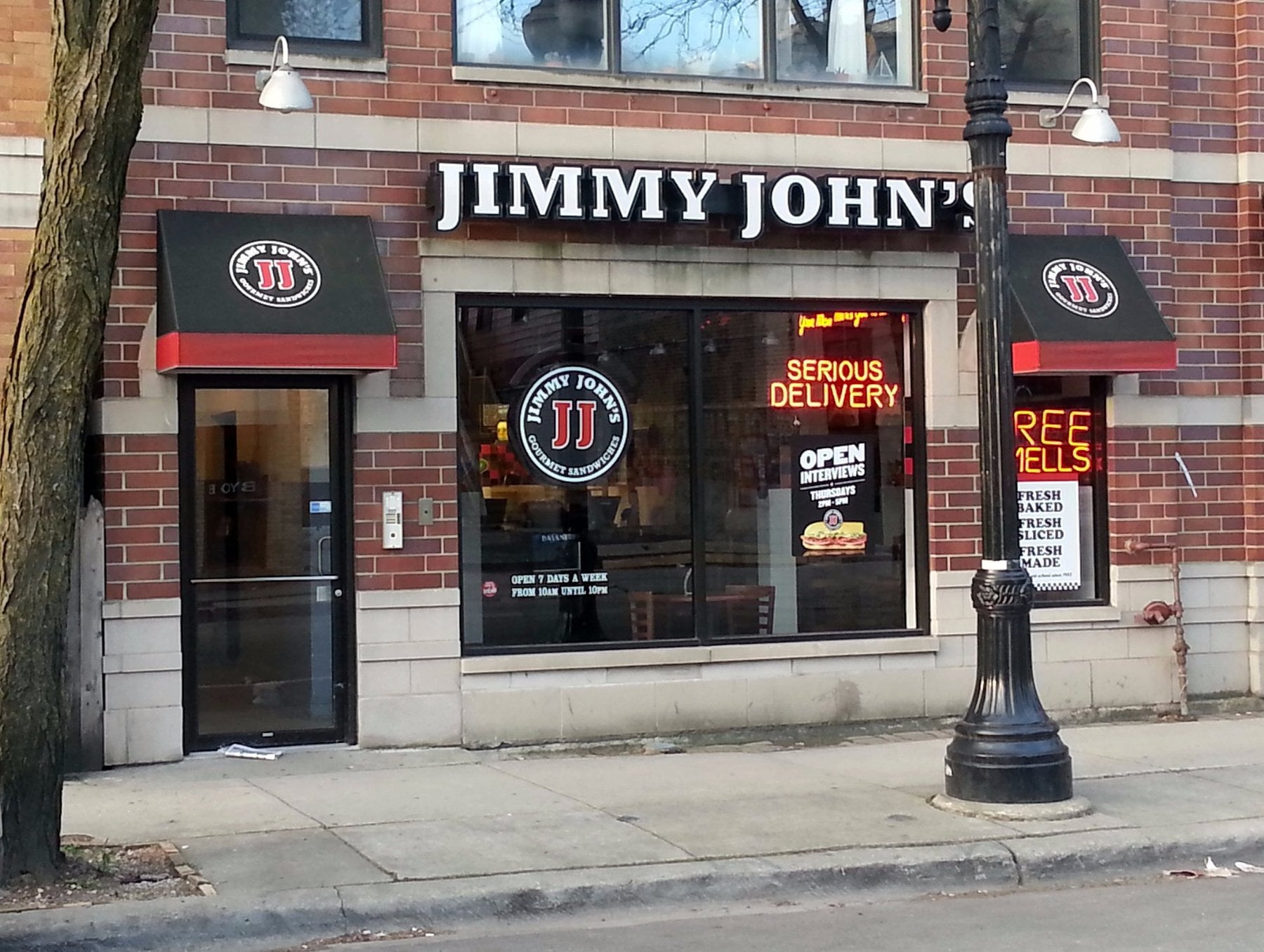 Jimmy John’s sandwich shop storefront with branded signs, a glass door, and window advertisements for delivery and open hours. A tree and streetlamp are in front.