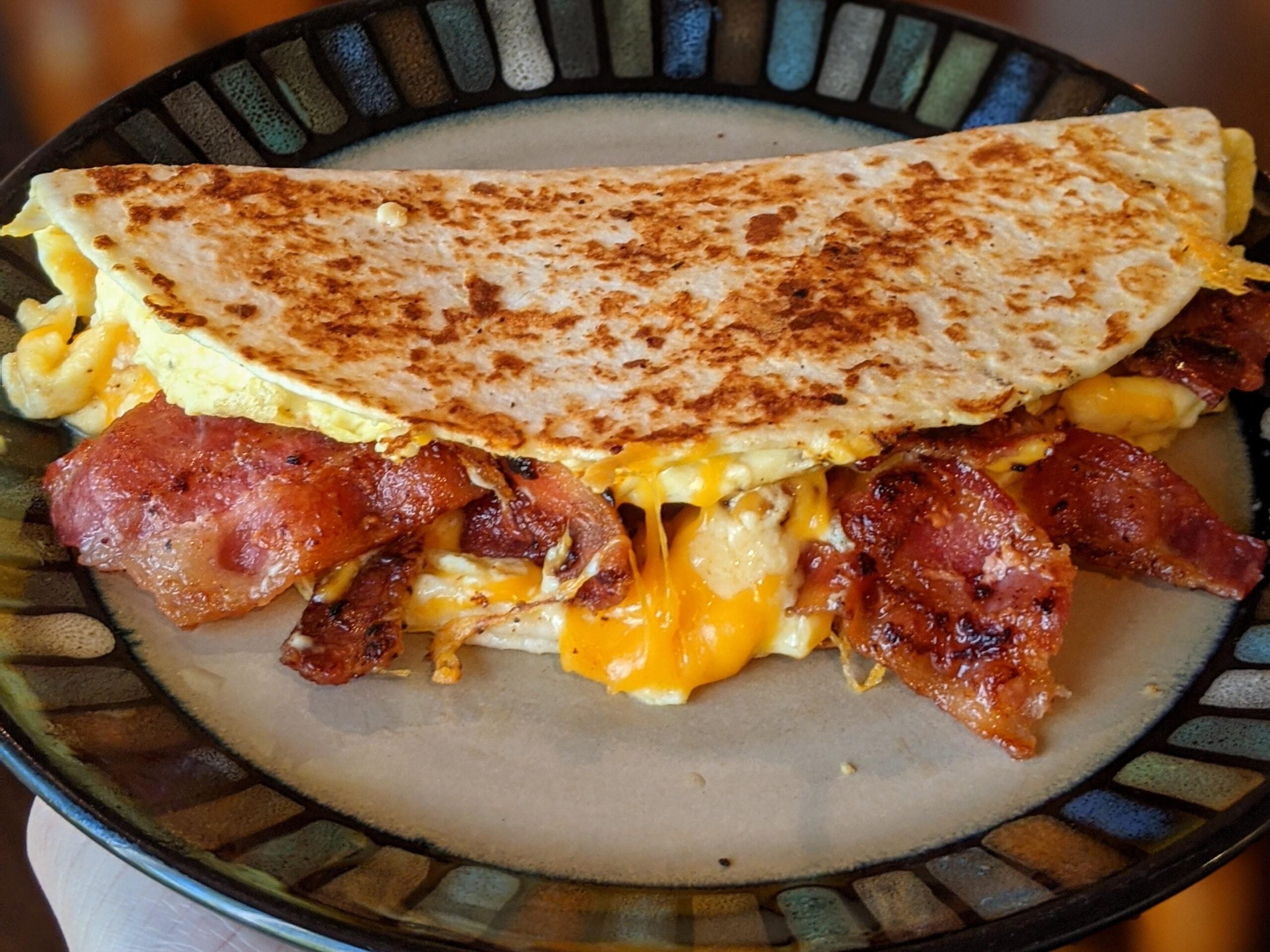 A hand holds a plate with a folded quesadilla filled with crispy bacon, scrambled eggs, and melted cheddar cheese.