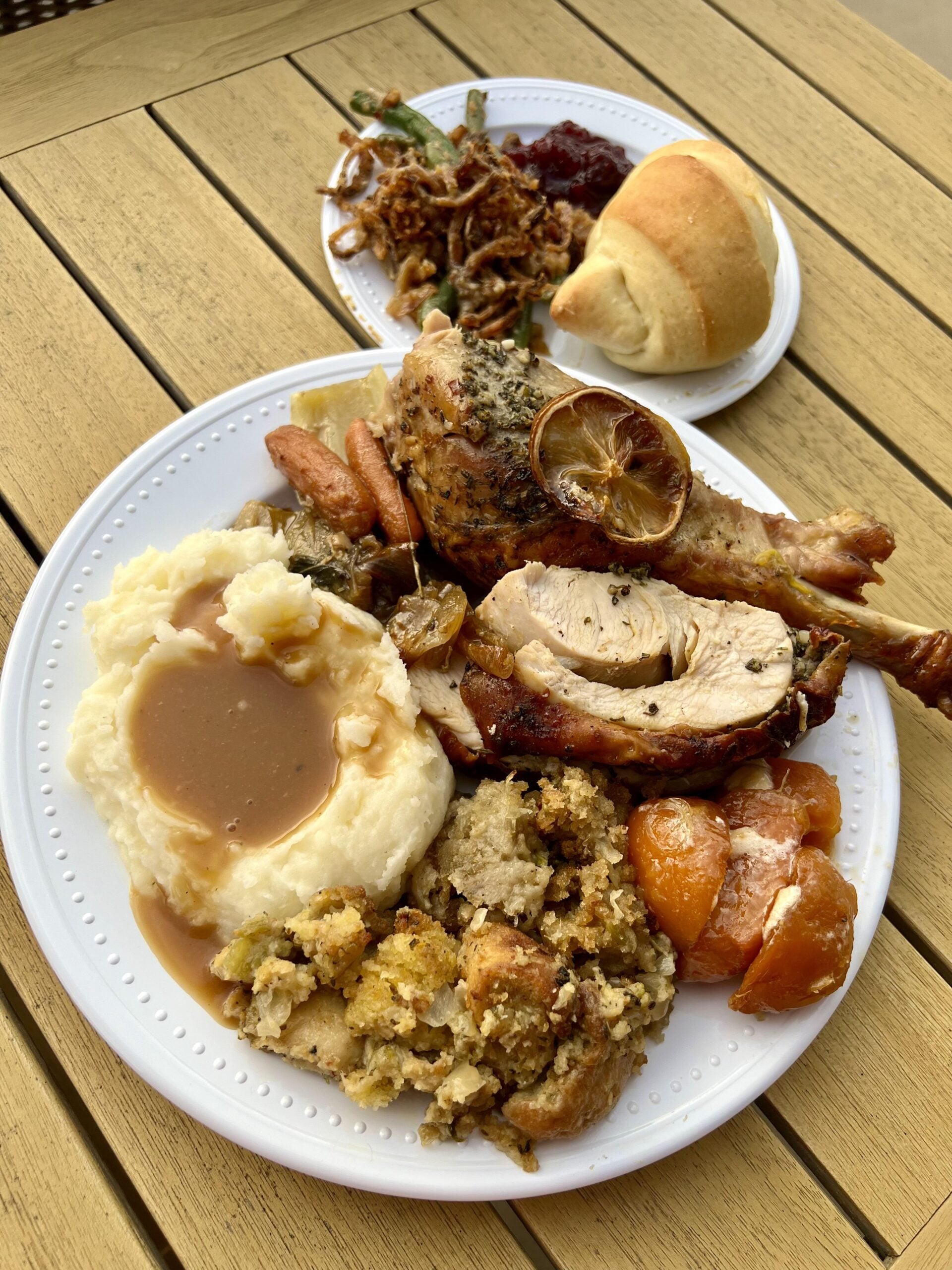 A plate with roasted turkey, stuffing, mashed potatoes with gravy, sweet potatoes, carrots, and green beans, next to a roll and cranberry sauce on a wooden table.