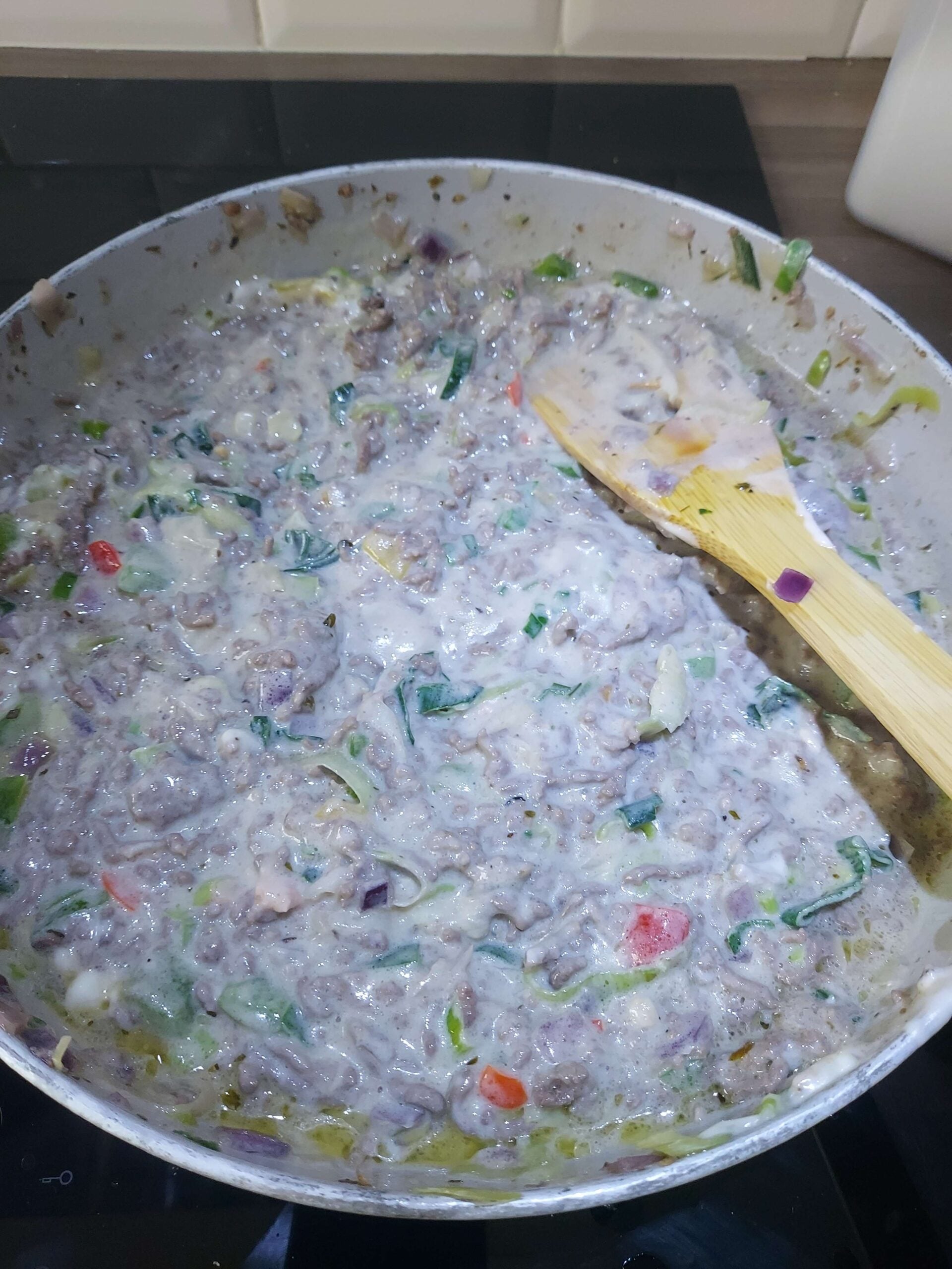A frying pan with a creamy, mixed vegetable and meat dish being stirred with a wooden spatula. The dish includes onions, red peppers, green vegetables, and chunks of meat.
