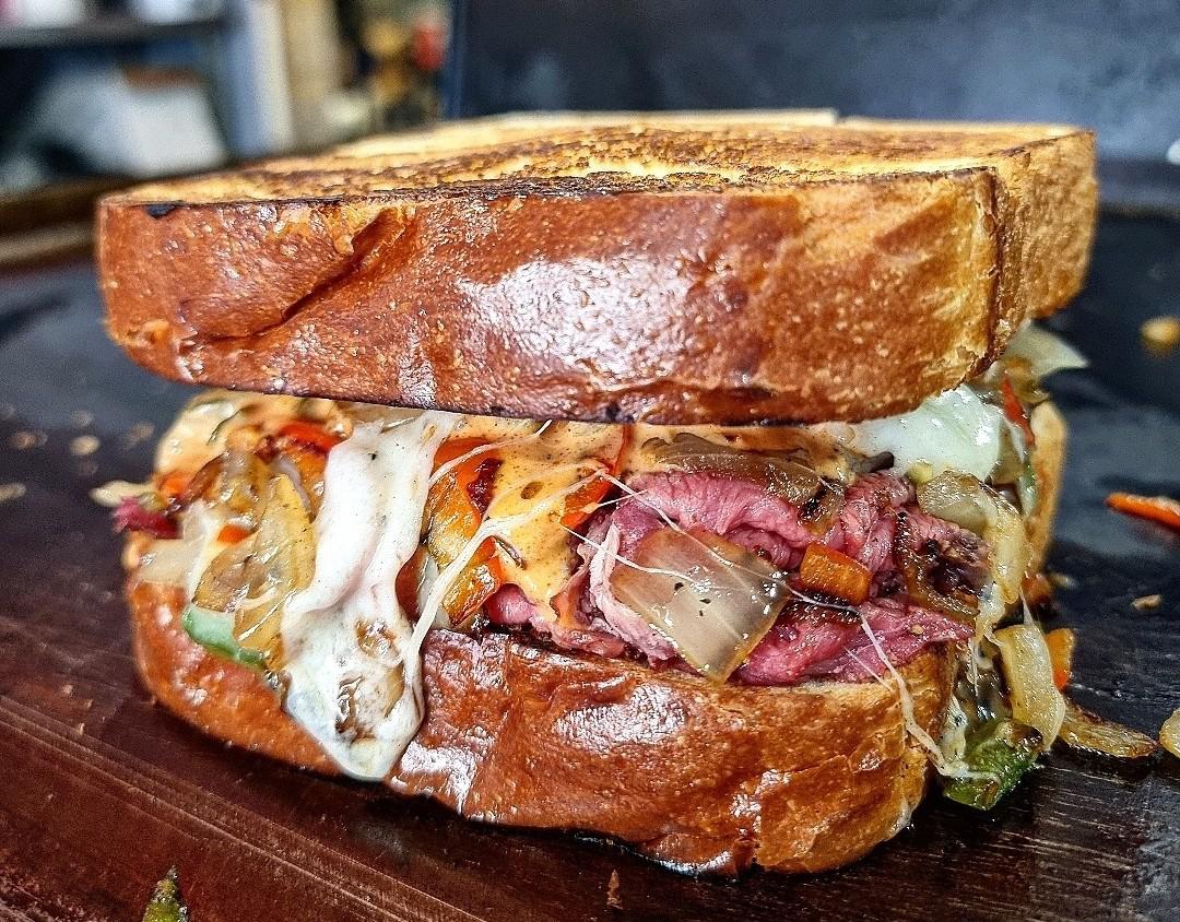 Close-up of a grilled sandwich with melted cheese, sliced beef, sautéed onions, and vegetables on thick toasted bread, resting on a wooden surface.