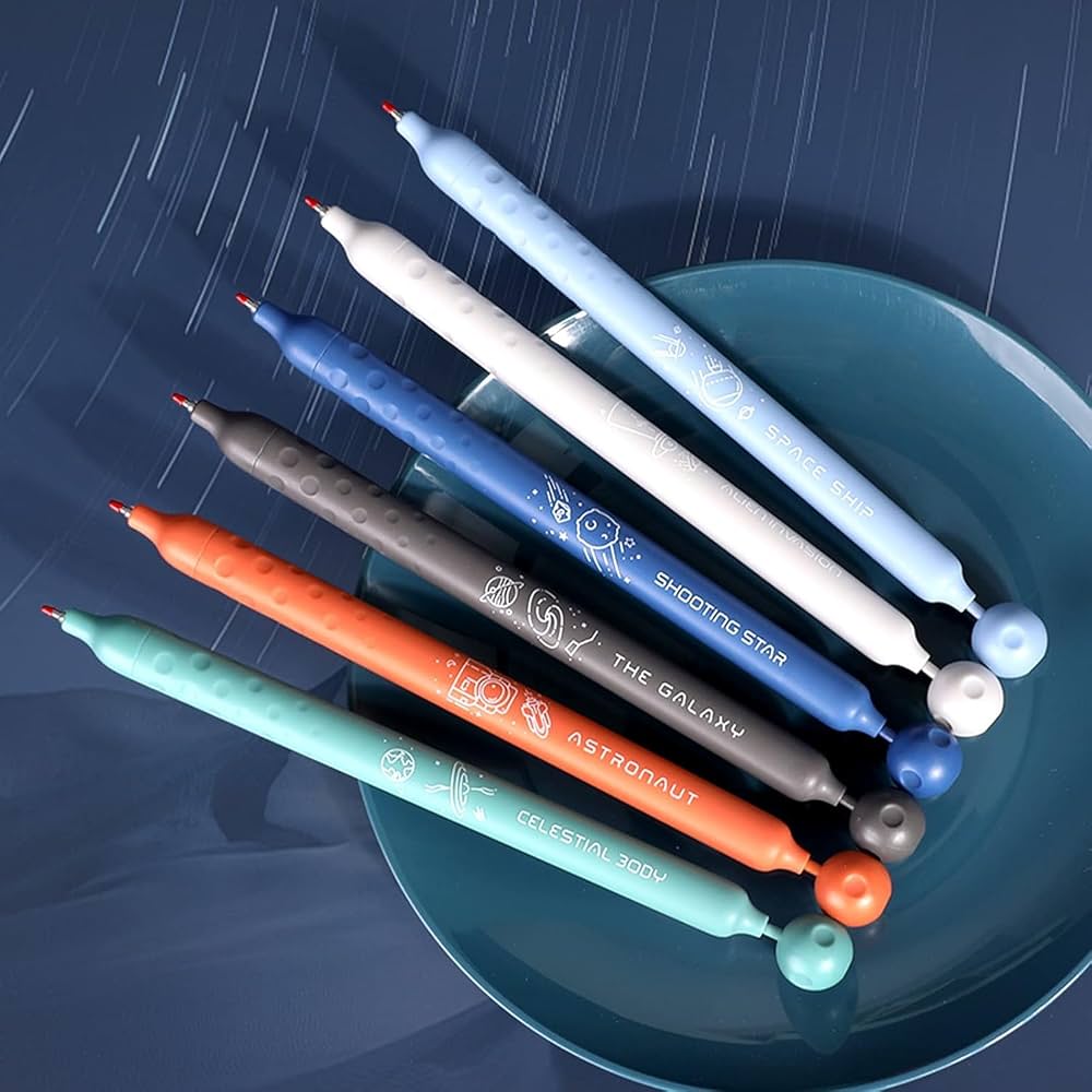 Seven colorful pens with space-themed designs and labels like “SPACE SHIP,” “SHOOTING STAR,” and “ASTRONAUT” are arranged on a blue plate against a dark blue, starry background.