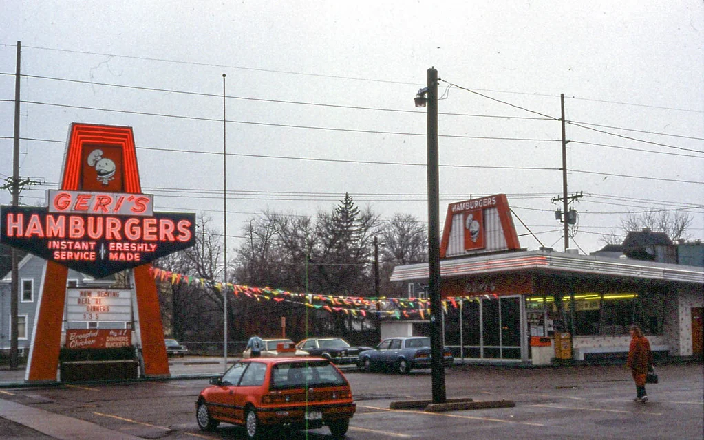 A vintage fast-food restaurant with a large neon sign reading “Geri’s Hamburgers.” A red car is parked outside, and colorful flags are strung across the lot. The sky is overcast and trees without leaves are in the background.
