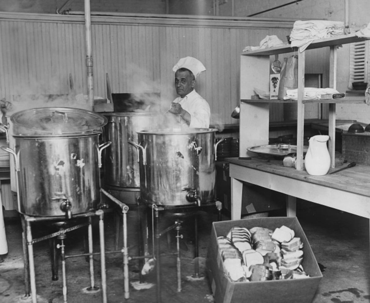 A chef in a white uniform stirs a large pot in an industrial kitchen, surrounded by steaming pots, shelves, dishes, and a box of bread slices in the foreground—perhaps preparing meals with historical significance that connect tradition and taste.