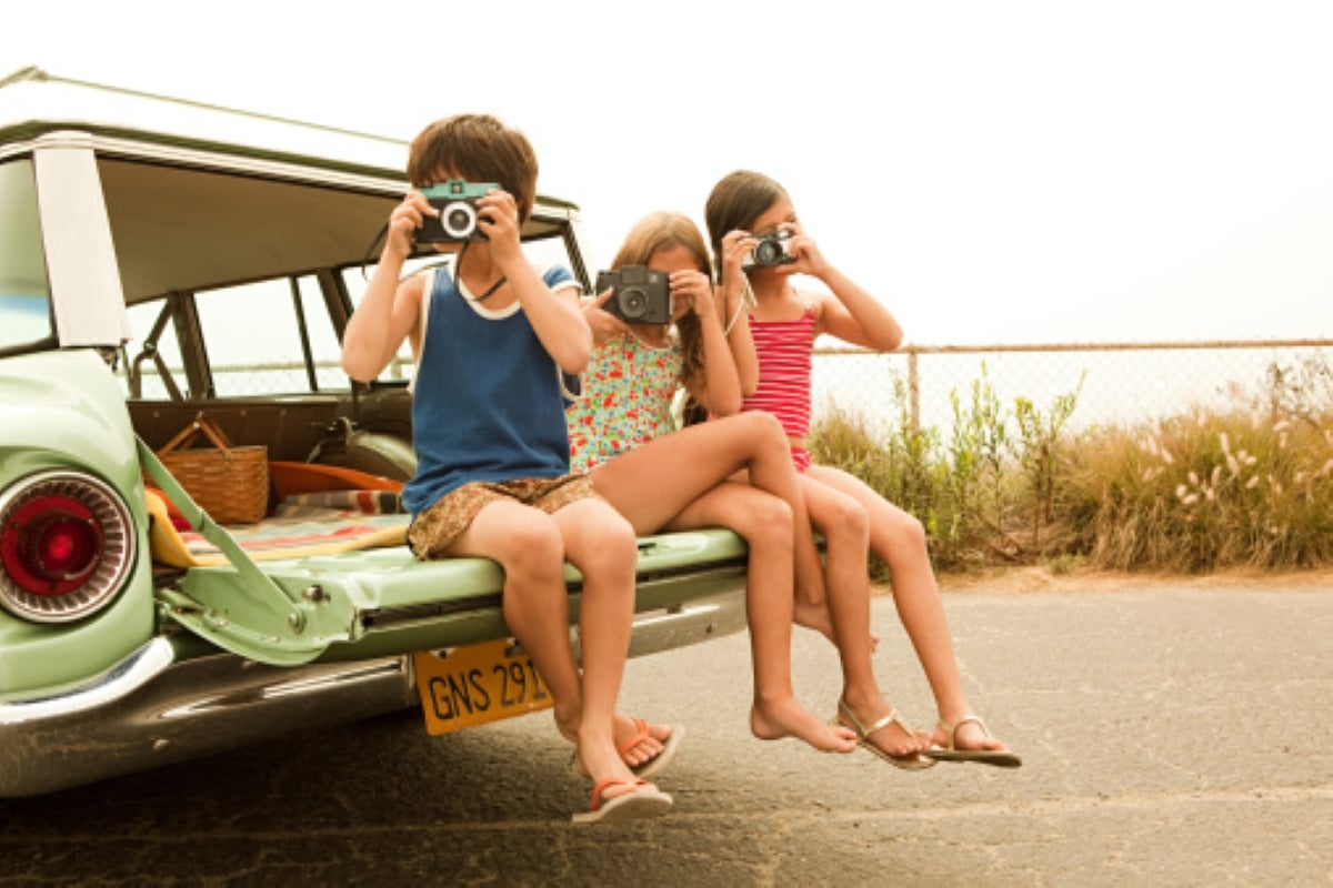 Three children sit on the open tailgate of a green car, each holding a camera and taking photos—capturing special moments from family road trips. They are dressed in summer clothes, and there is a picnic basket in the car. A fence and greenery are visible in the background.
