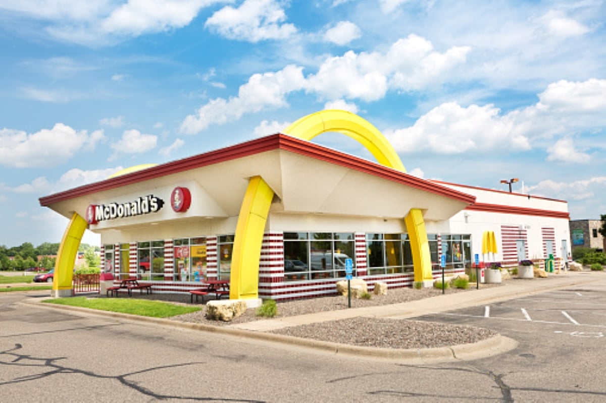 A McDonald’s restaurant with yellow arches, red accents, and striped decor stands out on a sunny day beneath a blue sky dotted with clouds. Perfect for family road trips, the location features empty parking spaces and inviting picnic tables outside.