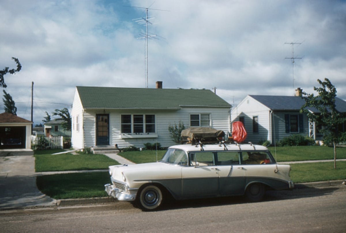 A vintage station wagon, perfect for family road trips, with luggage on the roof is parked in front of a small, single-story house with a green roof and well-kept lawn under a partly cloudy sky.