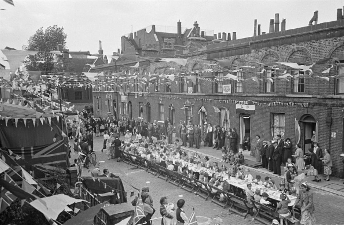 Black-and-white photo of a street party with rows of tables set for meals with historical significance, many people seated and standing, and bunting strung overhead in a residential area with brick houses.