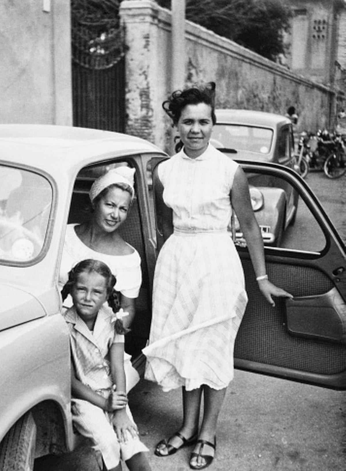 Three women pose next to a car on a city street, capturing the spirit of family road trips; one sits in the driver’s seat, another stands by the open car door, while a young girl perches on the car’s edge. Other cars and bicycles are visible in the background.