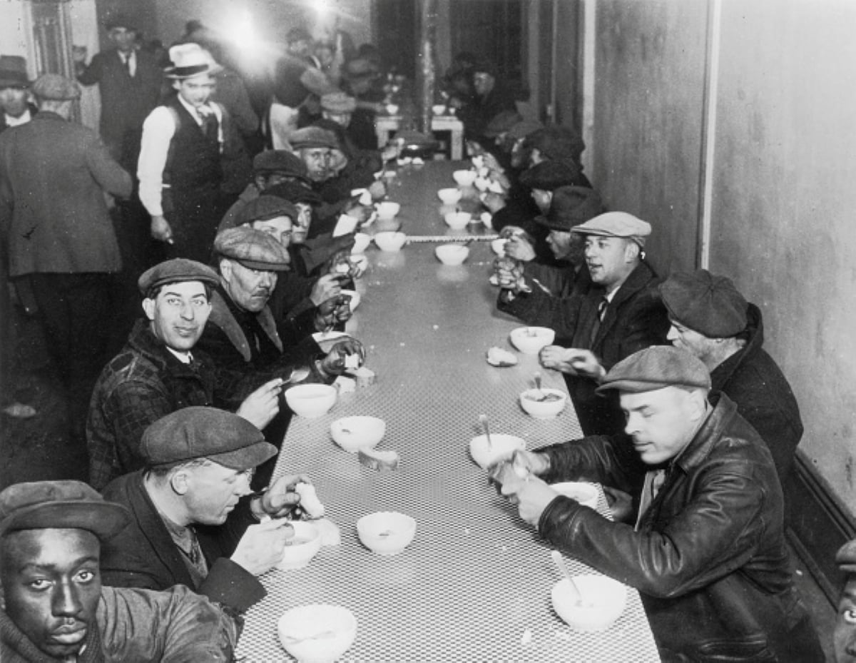 Black-and-white photo of men in caps eating at a long table in a soup kitchen, possibly during the Great Depression. This scene of meals with historical significance shows people seated closely, with bowls and spoons, some looking toward the camera.