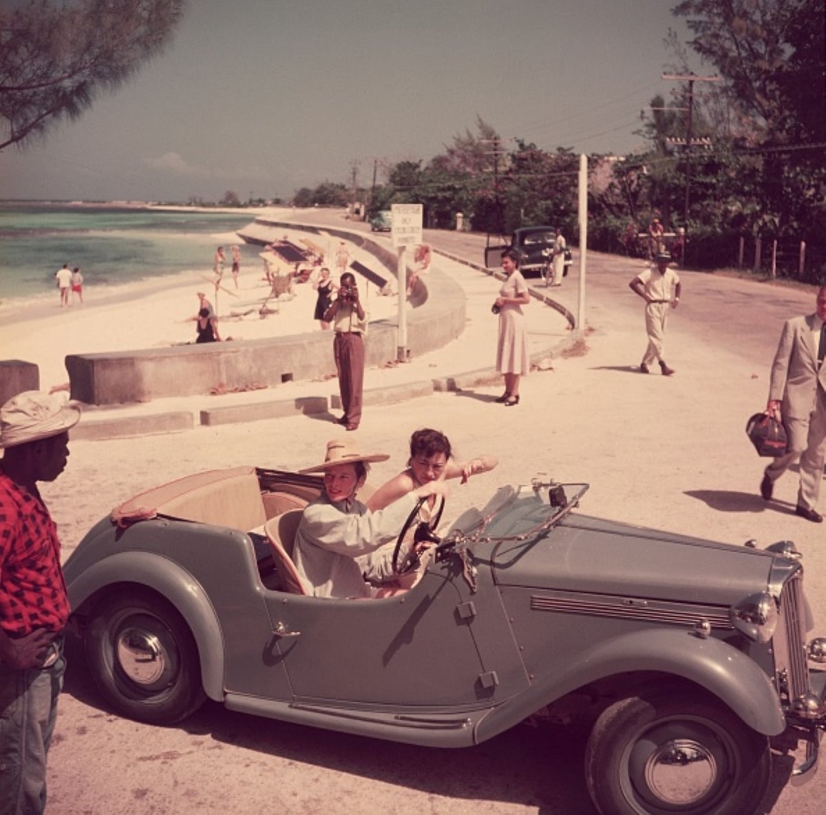 Two women in a vintage convertible smile near a beachside road, capturing the carefree spirit of family road trips; people walk and stand along the shore, with trees and the ocean in the background under a sunny sky.