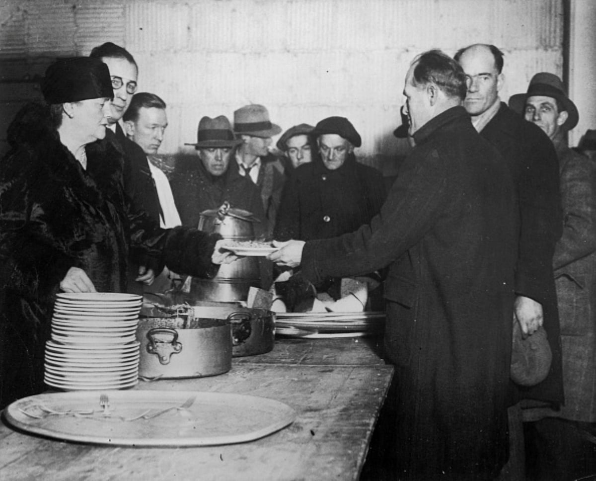 A group of people wait in line to receive food at a soup kitchen, partaking in meals with historical significance. A woman serves a man a meal while others stand nearby, holding plates. The scene appears to be from the early 20th century.