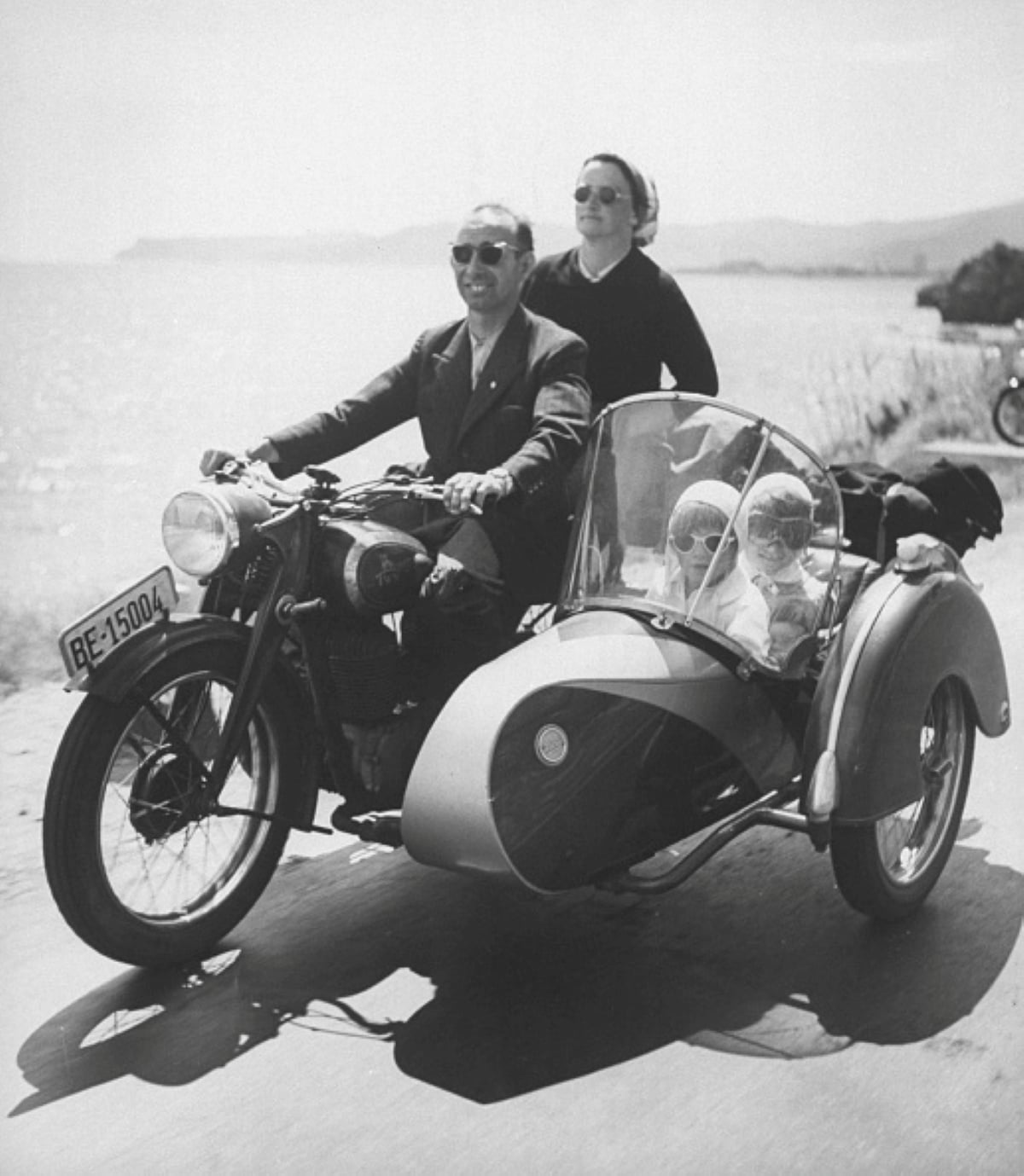 A man drives a motorcycle with a sidecar along a waterfront road, embodying the spirit of family road trips. A woman rides behind him and two children sit in the sidecar, all wearing sunglasses and smiling. It is a sunny day.