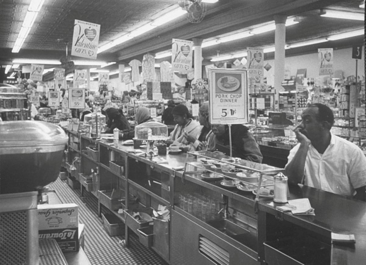 Black and white photo of people seated at a busy diner counter with food, under signs advertising pork chop dinners for 59 cents. Shelves of goods are visible in the background, capturing meals with historical significance from a bygone era.