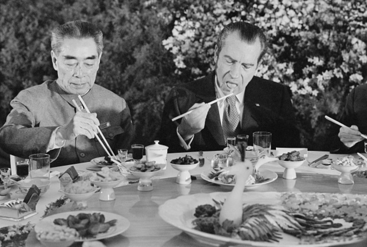 Two men in suits eat at a round table set with various Chinese dishes, perhaps enjoying meals with historical significance. One man uses chopsticks skillfully while the other, wearing a suit and tie, sticks out his tongue in concentration as he attempts to eat with chopsticks.