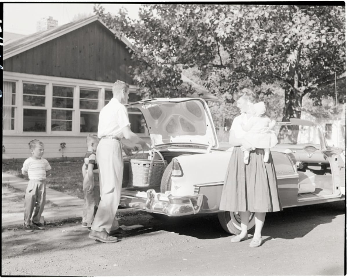 A man loads picnic baskets into a car trunk while a woman holding a baby and a young child stand nearby in front of a house, suggesting the start of one of their cherished family road trips.