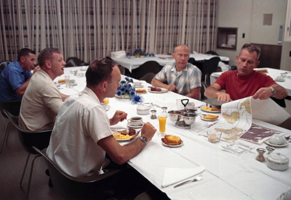 Five men sit at a table with breakfast foods, drinks, and tableware; one man in a red shirt examines a map while others look on. The room has patterned curtains and a simple, functional decor, suggesting an atmosphere where meals with historical significance might be shared.