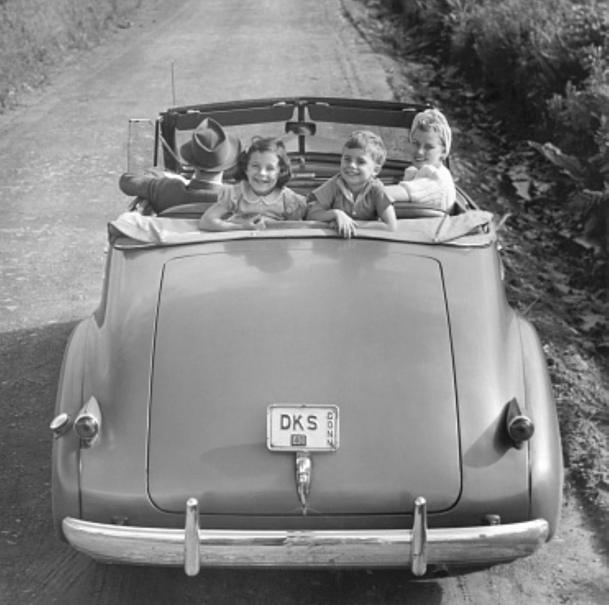 Three smiling children sit in the back of a vintage convertible car on a dirt road, enjoying one of their family road trips, with an adult driving. The car has a “DKS” license plate and trees line the roadside.