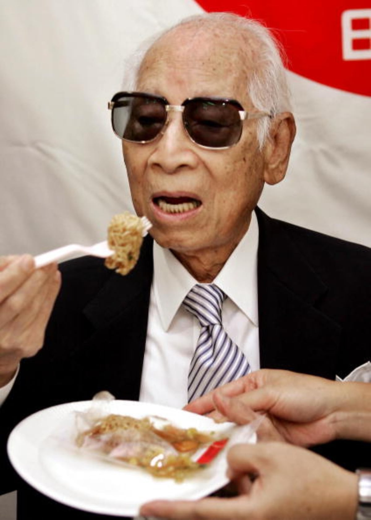 An elderly man in a suit and sunglasses is about to eat food with a plastic fork from a paper plate, perhaps enjoying one of those meals with historical significance. A hand holds the plate for him. A red and white background is visible behind him.