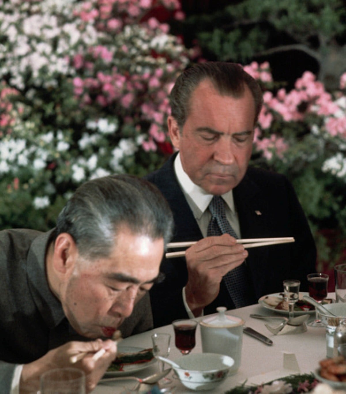 Two men sit at an outdoor table with flowers in the background, enjoying meals with historical significance as they eat with chopsticks. One man is bowing over his bowl while the other looks at his food intently, savoring each bite. Various dishes and drinks are on the table, adding to the rich culinary experience.