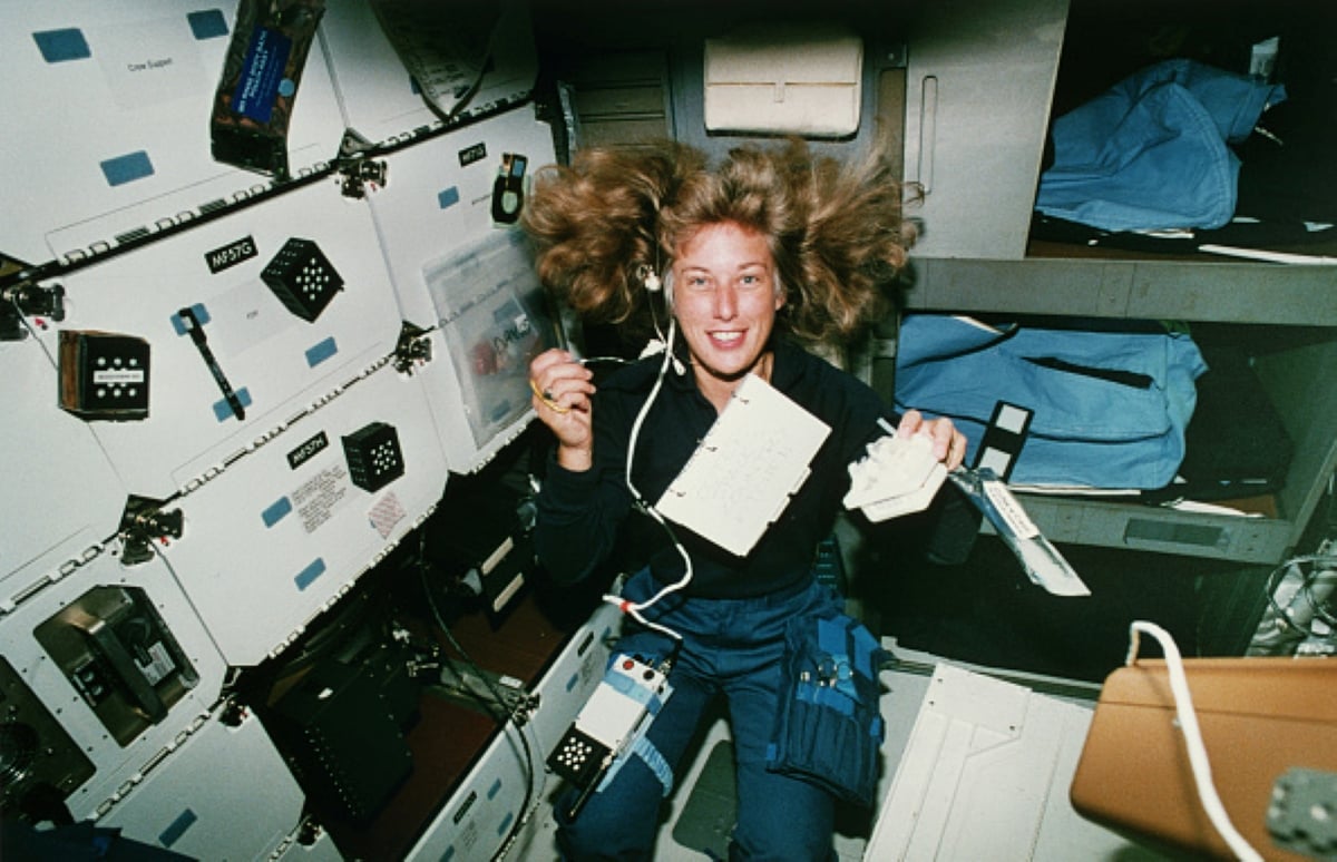 An astronaut with floating hair smiles while holding tools and working inside a spacecraft surrounded by equipment, storage compartments, and specially packed meals with historical significance.