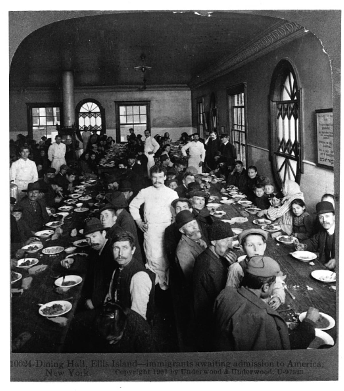 Black-and-white photo of immigrants seated at long tables in a crowded dining hall on Ellis Island, New York, awaiting admission to America. Dishes and cups are on the tables; several people face the camera, sharing meals with historical significance as they begin a new chapter in their lives.