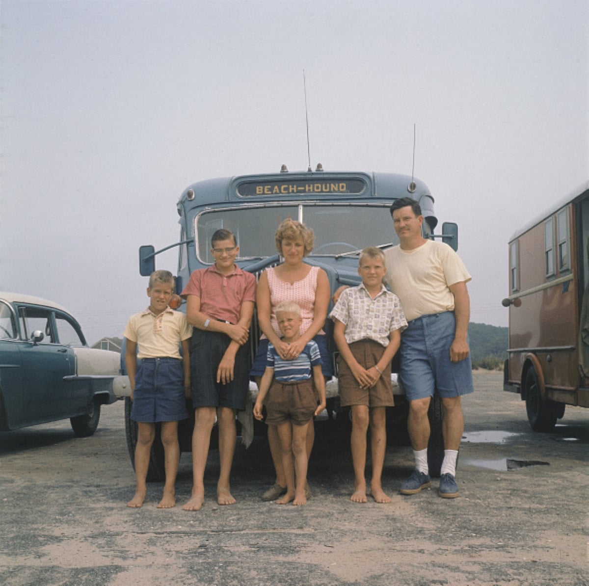 A family of six, barefoot and smiling, stands in front of a blue bus labeled “Beach-Hound” in a parking lot—two cars parked on each side—capturing the joy and togetherness found on family road trips.