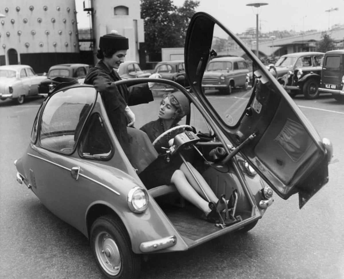 Two women sit in a small, three-wheeled car with a front-opening door, parked among vintage cars in a city lot. Reminiscent of classic family road trips, one woman is entering while the other sits inside, smiling.