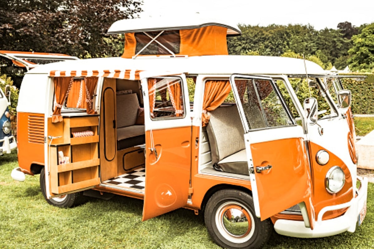 Vintage orange and white camper van with open doors, showing a retro interior with checkered flooring, orange curtains, and a pop-up roof—perfect for family road trips—parked on grass with trees and greenery in the background.