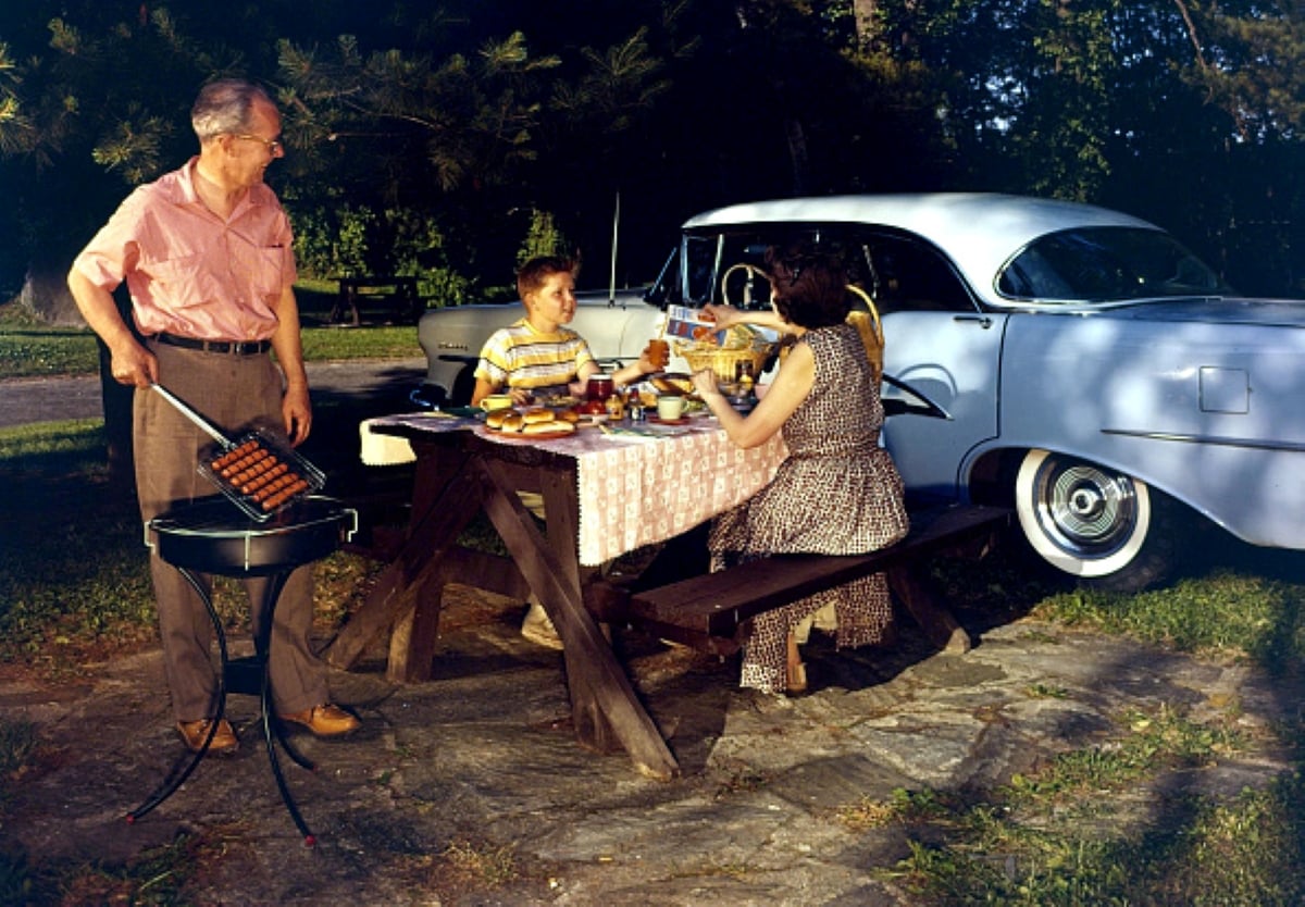 A man grills food beside a picnic table where a woman and a boy are eating, capturing the joyful spirit of family road trips. A vintage car is parked nearby, and the scene takes place outdoors on a sunny day.