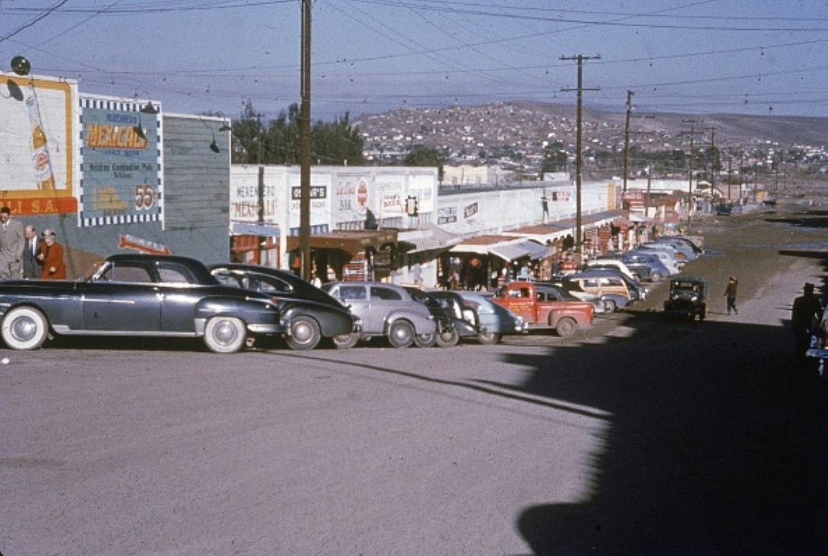A 1950s street scene shows vintage cars, reminiscent of family road trips, parked along storefronts with colorful signs, people on the sidewalk, and hills with buildings in the background under a clear sky.