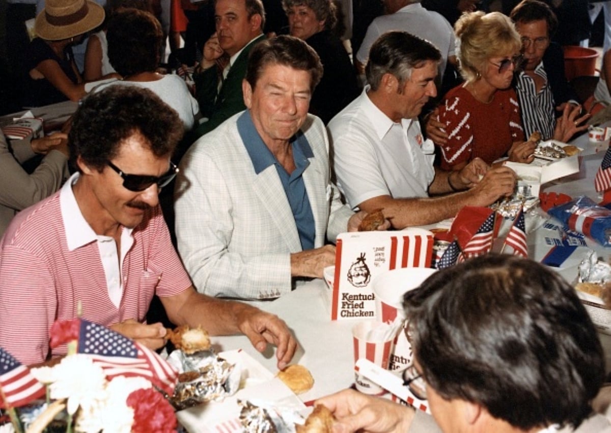 A group of people sit at a table eating Kentucky Fried Chicken, with American flags decorating the table. The setting appears festive and social, possibly a celebration or public event—one that highlights meals with historical significance in American culture.