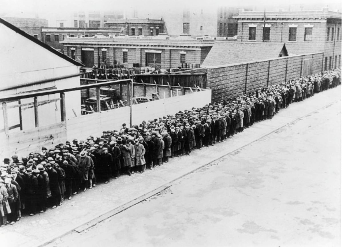 A long line of people dressed in coats and hats stand on a city sidewalk beside a wooden fence and buildings, likely waiting for meals with historical significance or assistance during the Great Depression.