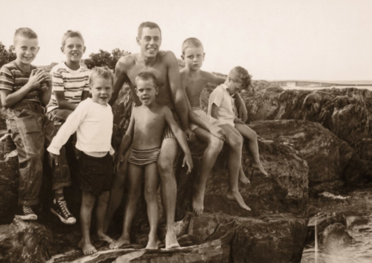 Six boys and one man sit and stand on rocky terrain near water, smiling—capturing the carefree spirit often found during family road trips. Some are barefoot, and some wear striped shirts or swimsuits. The image is in sepia, giving it a vintage look. Trees and water are visible in the background.