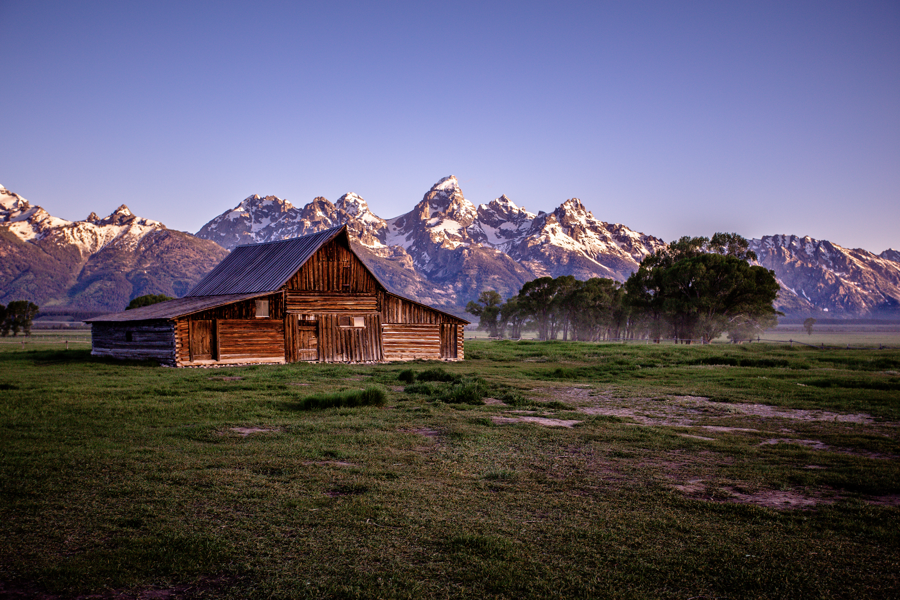 A rustic wooden barn stands on green grass with snow-capped mountains and a clear purple sky in the background.