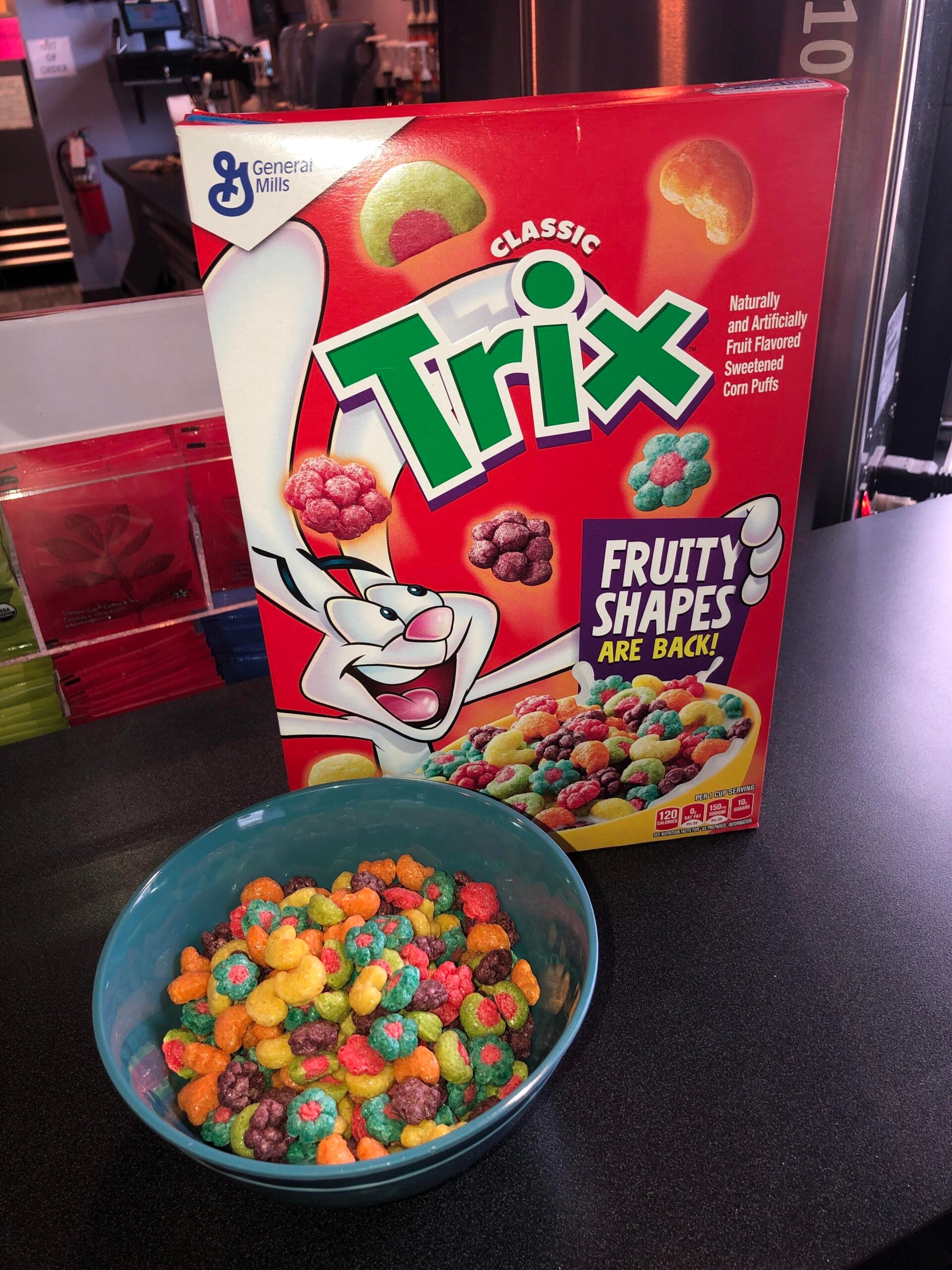 A box of Trix cereal with the Trix rabbit mascot is behind a bowl filled with colorful, fruit-shaped cereal pieces on a countertop.