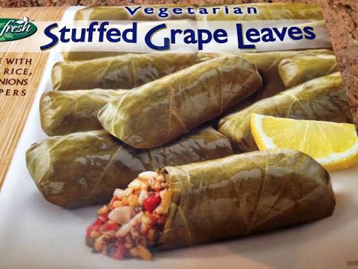A close-up of vegetarian stuffed grape leaves, some whole and one cut open to show a filling of rice and vegetables, with a lemon wedge on the side.
