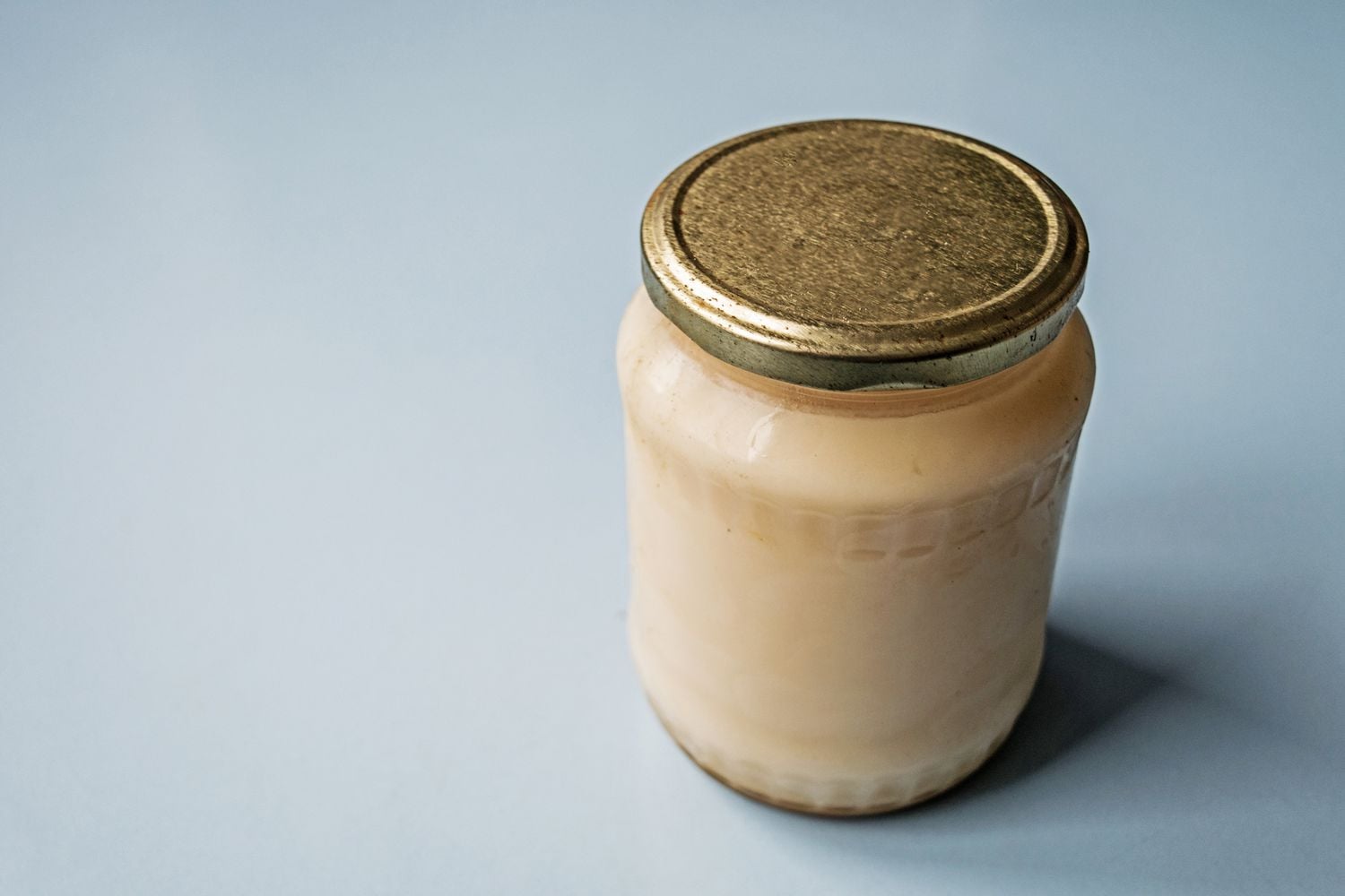 A small glass jar with a golden metal lid, filled with a creamy white substance, sits on a light blue surface.