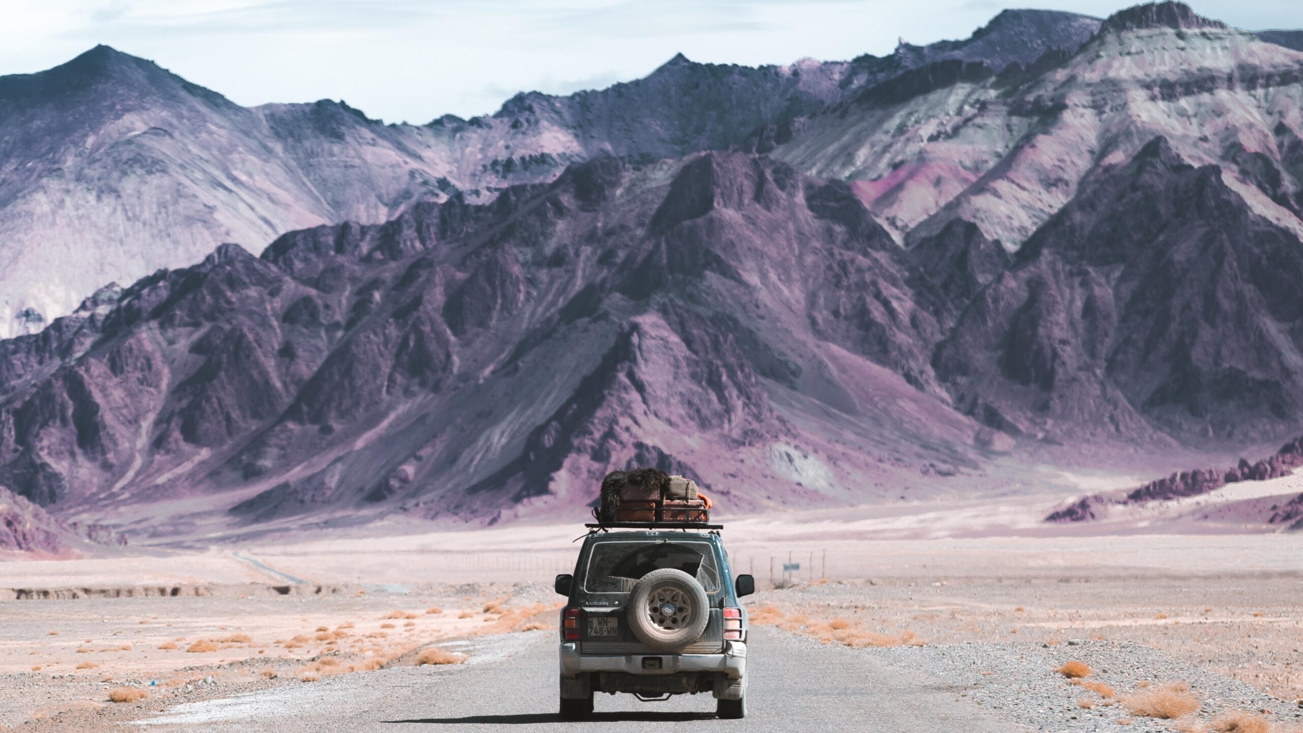 A car loaded with luggage drives on a deserted road toward rugged, purple-hued mountains under a partly cloudy sky in a barren landscape.