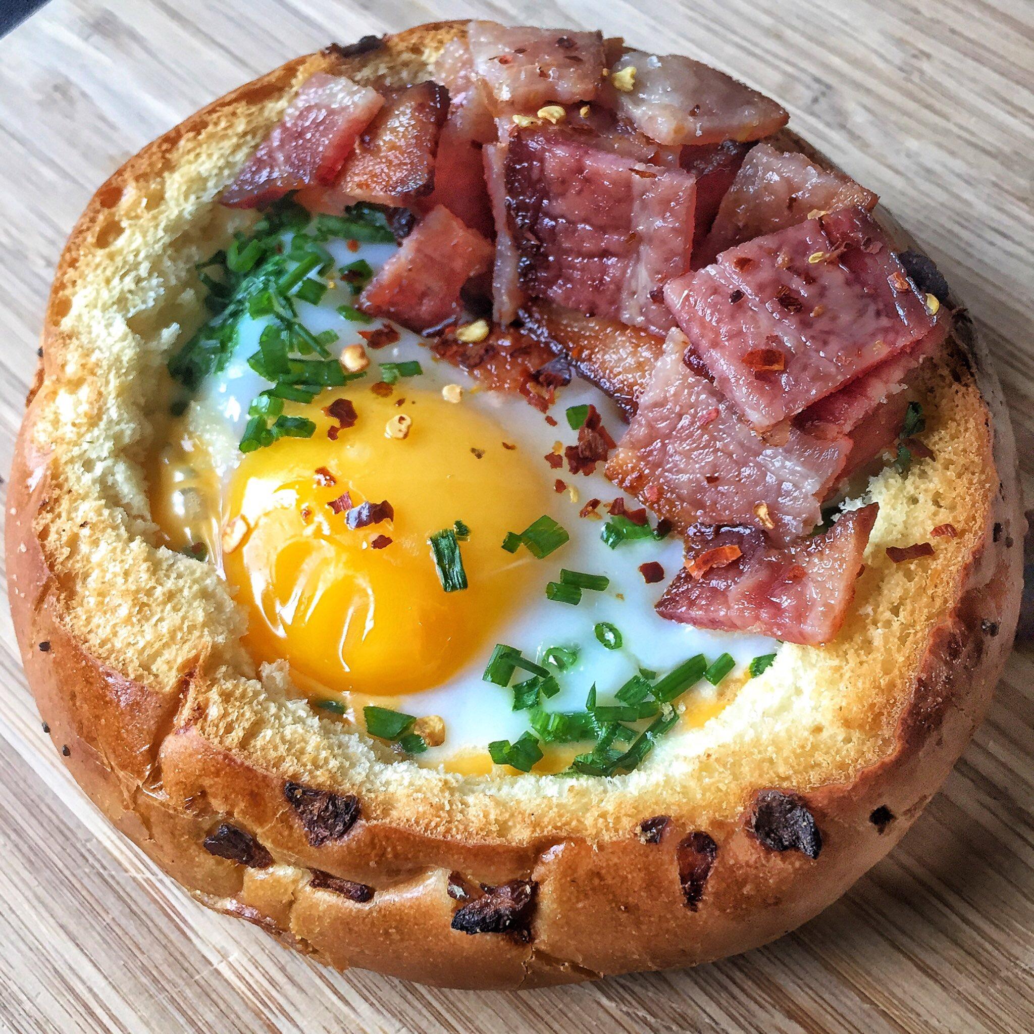 A bread roll filled with a baked egg, chopped ham, chives, and red pepper flakes, sitting on a wooden surface.