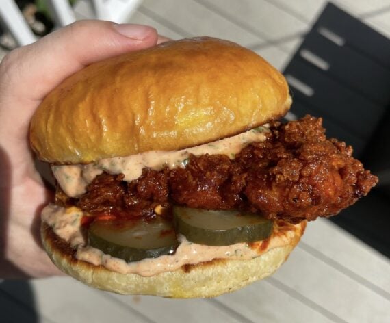 A hand holds a fried chicken sandwich with pickles and creamy sauce on a shiny brioche bun, outdoors in sunlight.