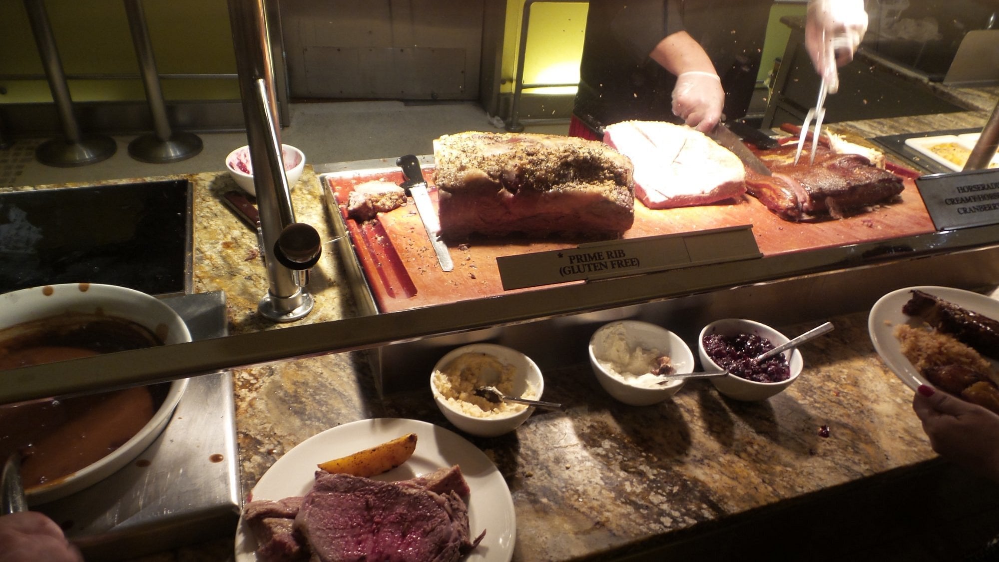 A buffet carving station with a chef slicing prime rib. Plates with meat, sides, and sauces are visible in the foreground. A sign reads “Prime Rib Gluten Free.” The scene is well-lit and the counter is granite.